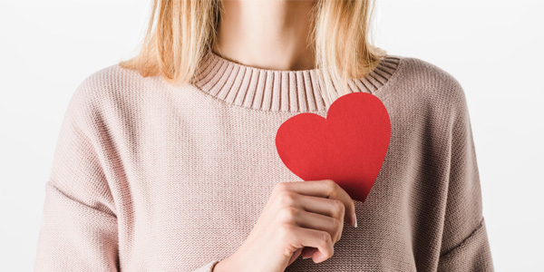 Person trägt einen hellrosa Strickpullover und hält ein rotes Papierherz vor die Brust.
